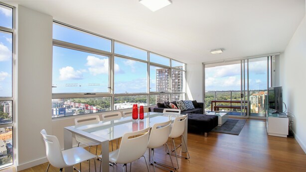 A modern living room with a large glass table and white chairs, a black sofa, and a TV. - Modern and Bright 3BR Executive Apartment in Zetland with Views (Zetland)