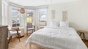 A bedroom with a large bed, two chairs, a small table, a ceiling fan, and a view of the outside through large windows. - Romantic Queen Suite 1 Block to Beach & Asbury | Victorian Charm | 1000 Mbps ... (Neptune Township)
