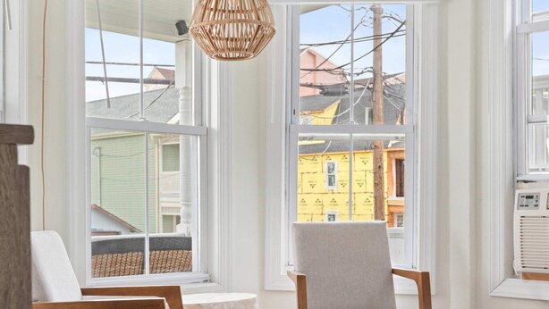 A bedroom with a bed, two chairs, a hanging light fixture, and large windows with a view of buildings. - Romantic Queen Suite 1 Block to Beach & Asbury | Victorian Charm | 1000 Mbps ... (Neptune Township)