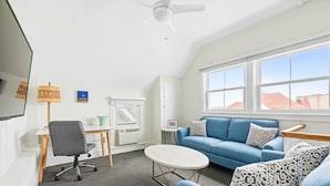 Ein modernes Zimmer mit einem blauen Sofa, einem kleinen runden Tisch, einem grauen Bürostuhl, einem Deckenventilator und einem großen Fenster mit Blick auf Gebäude.