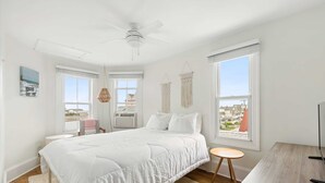 Une chambre à coucher avec un grand lit, un ventilateur de plafond, deux fenêtres, une petite table ronde et un meuble pour la télévision.
