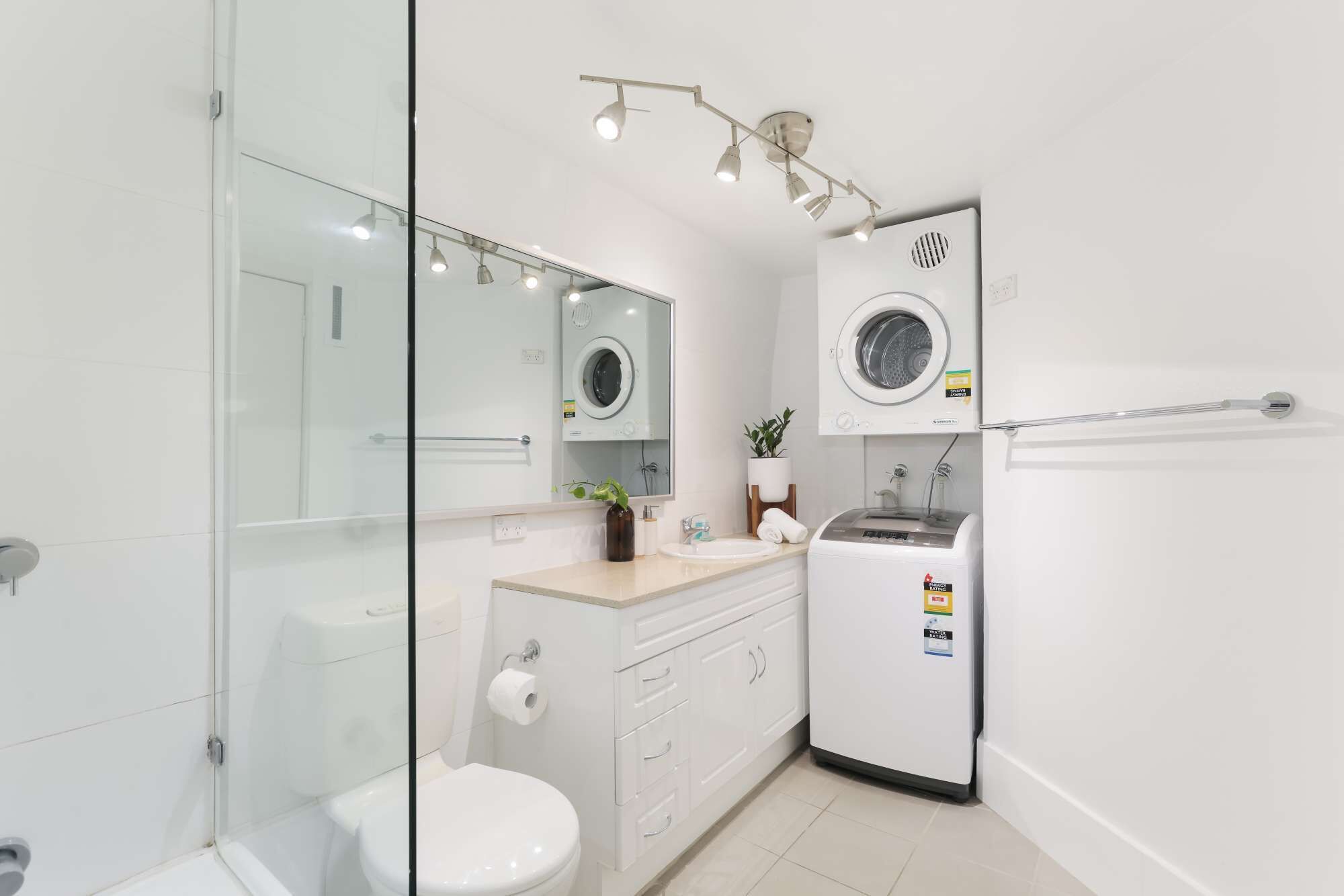 Ein modernes Badezimmer mit Duschkabine aus Glas, Toilette, Waschbecken mit Spiegel und integrierter Waschmaschine.