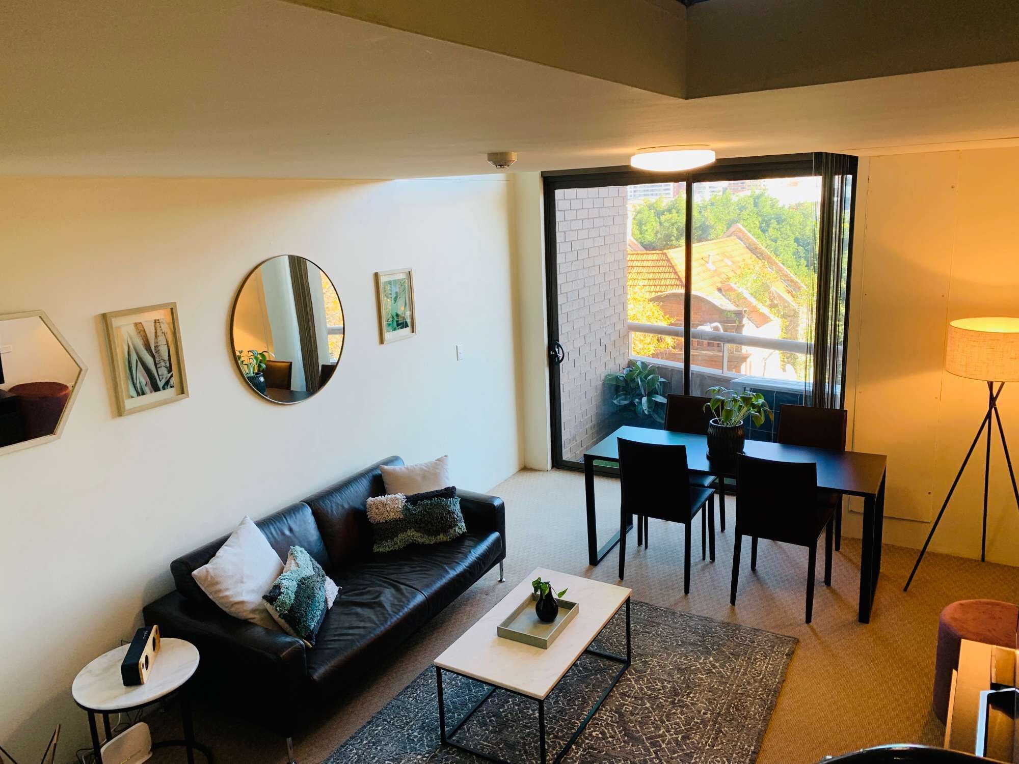 A modern living room with a black leather sofa, a coffee table, and a dining area with chairs and a table. There is a large window with a view of a brick building and greenery outside.