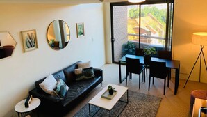 A modern living room with a black leather sofa, a coffee table, and a dining area with chairs and a table. There is a large window with a view of a brick building and greenery outside.