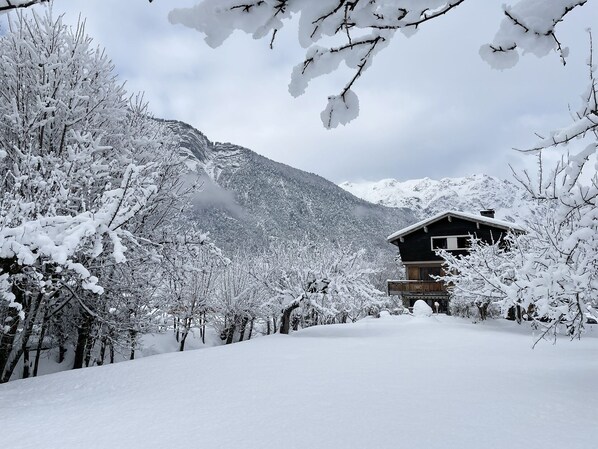 Una cabaña de madera con balcón, rodeada de árboles cubiertos de nieve y montañas al fondo.