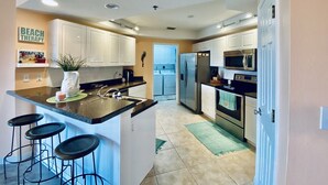 A modern kitchen with white cabinets, a stainless steel refrigerator, a microwave, and a sink. There are three black bar stools and a potted plant on the counter.