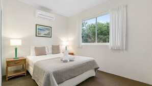 A hotel room with a bed, two bedside tables, a window with curtains, an air conditioning unit, and a framed picture on the wall.