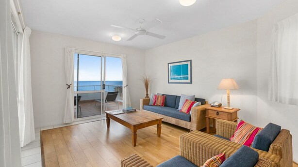 A spacious living room with a large window overlooking the sea, a ceiling fan, a sofa set, a coffee table, and a side table with a lamp.