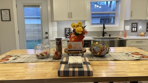 Une cuisine avec des meubles blancs, une table en bois avec des sets de table, un vase rempli de fleurs et une décoration sur le thème de Thanksgiving.