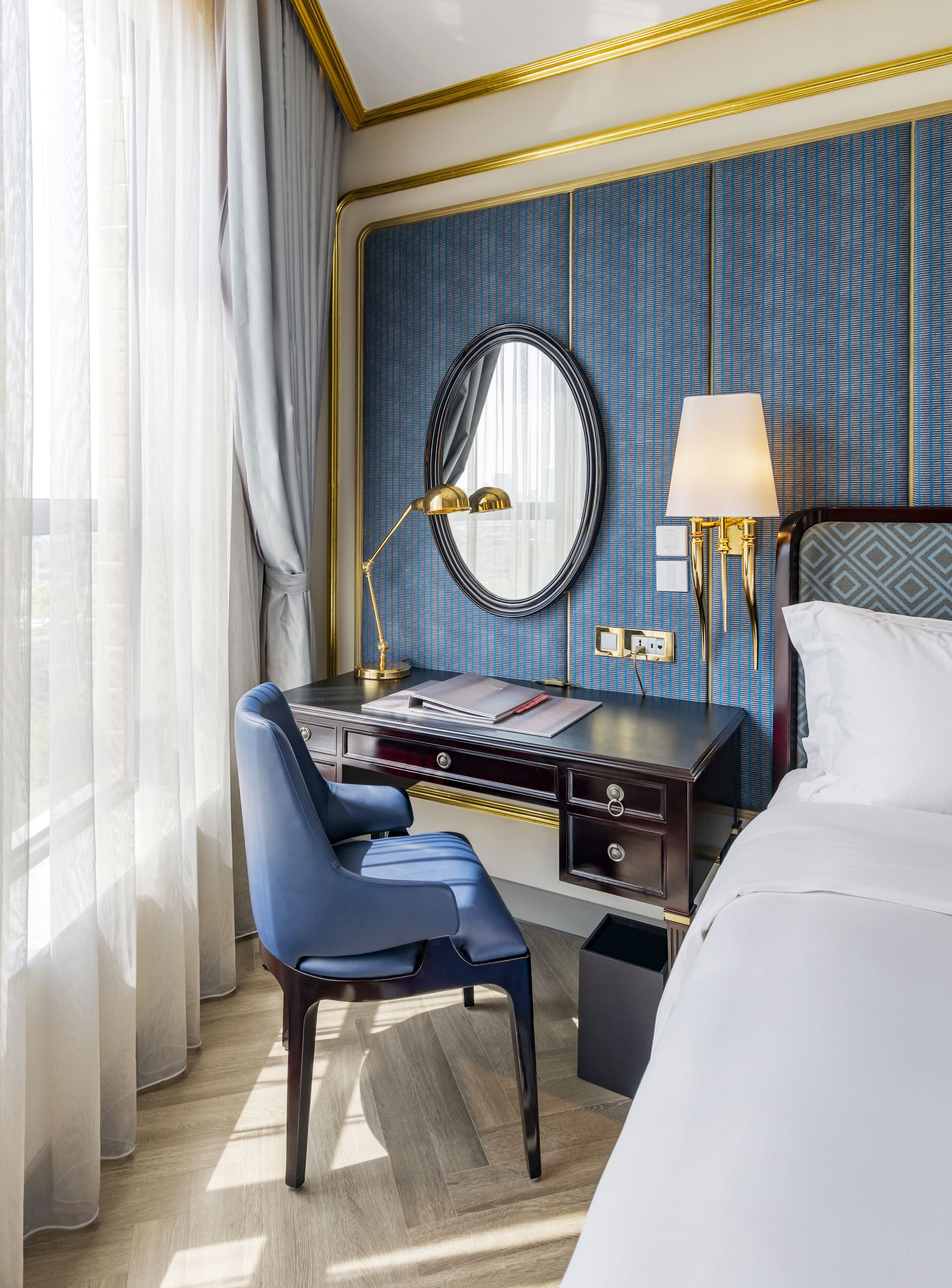 Une chambre d’hôtel comprenant un lit, un bureau avec une chaise, un miroir, une lampe et une fenêtre avec des rideaux.