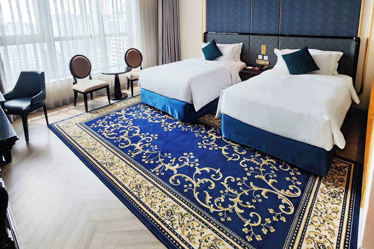 A hotel room with two beds, a large blue and gold patterned rug, a desk, and a chair.