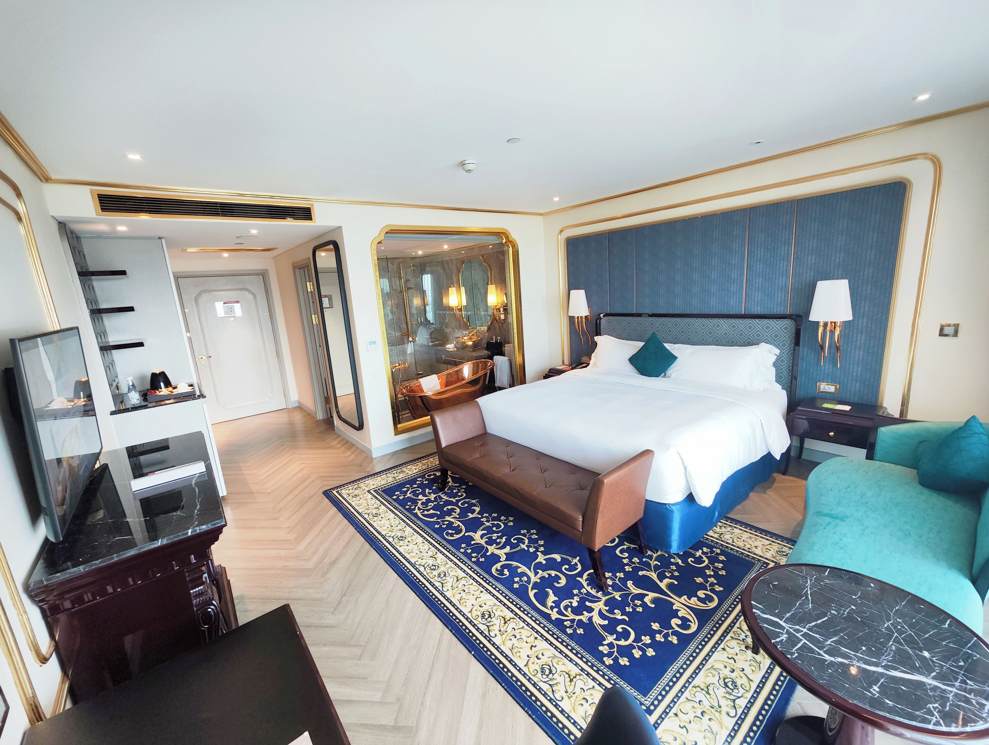 A hotel room with a large bed, a sitting area, a desk, a TV, and a bathroom with a bathtub.