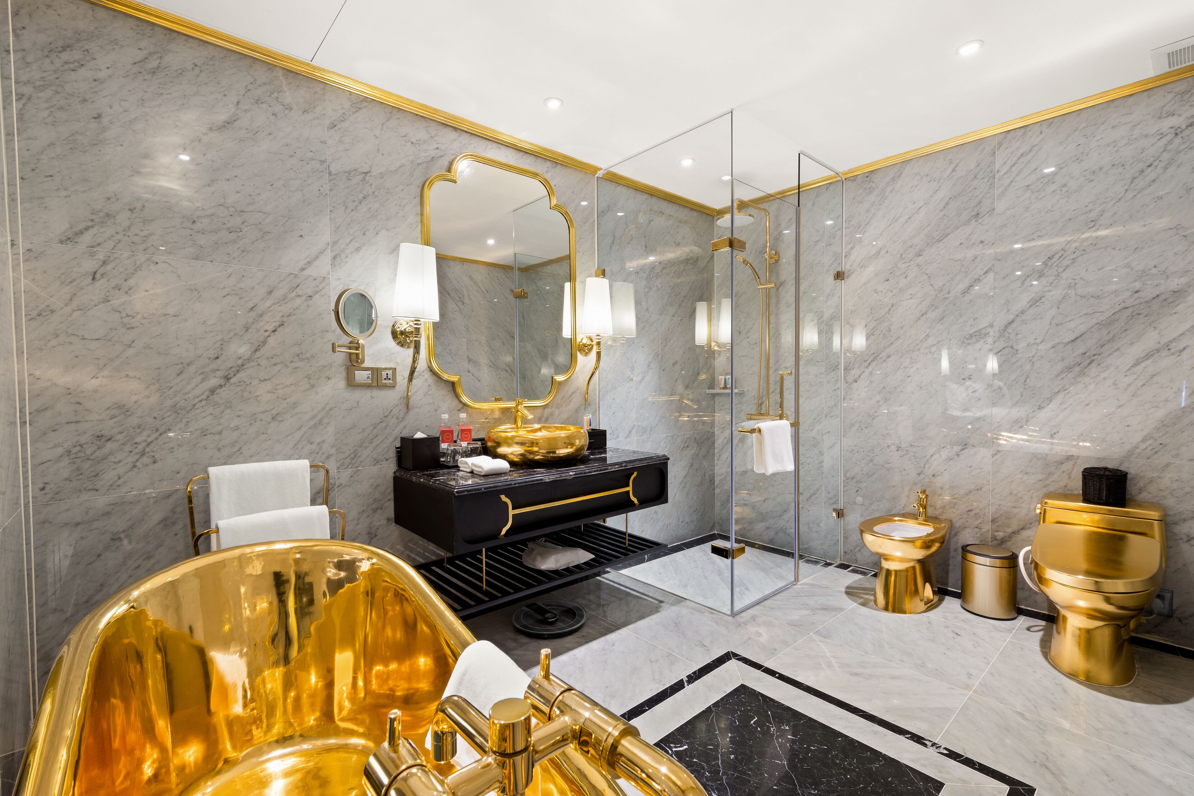Un baño lujoso con pared de mármol, una tina grande dorada, un tocador negro con detalles dorados y un inodoro dorado.
