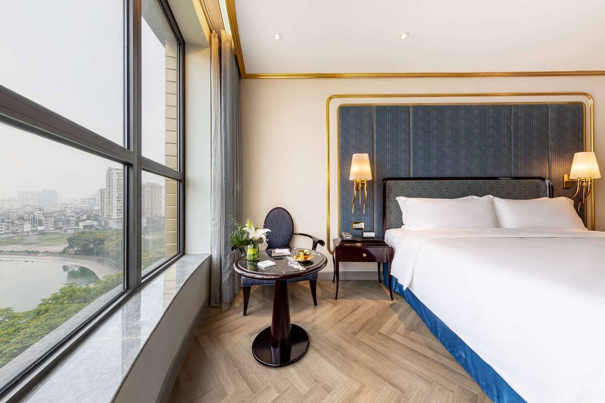 A hotel room with a large window, a bed, a round table with a vase, and two bedside lamps.