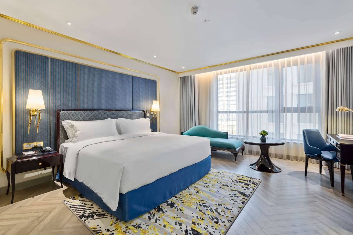 A modern hotel room with a large bed, a desk, a chair, a small table, and a window with curtains.
