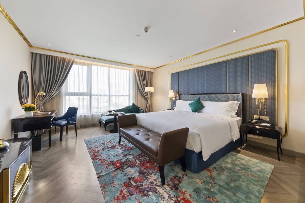 A hotel room with a large bed, a desk with a piano, a sofa, and a view of the city through large windows.