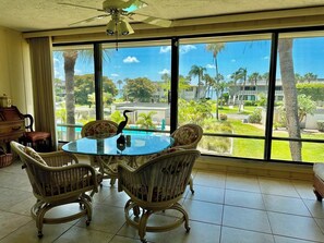 Quarto com mesa de vidro rodeada por cadeiras de vime, um ventilador de teto e vista para uma área residencial com palmeiras e casas.