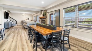 A spacious dining area with a wooden table and black chairs, a kitchen with a stone fireplace, and large windows with blinds. - Breathtaking Home! *In Town*Fire-Pits* Indoor Pool*Yard Games*Golf View* (Branson)