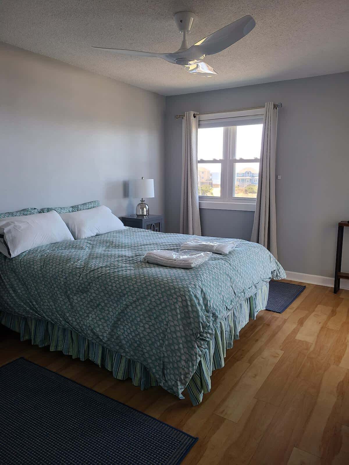 Een slaapkamer met een bed, een plafondventilator, een raam met gordijnen, een nachtkastje en uitzicht op gebouwen.