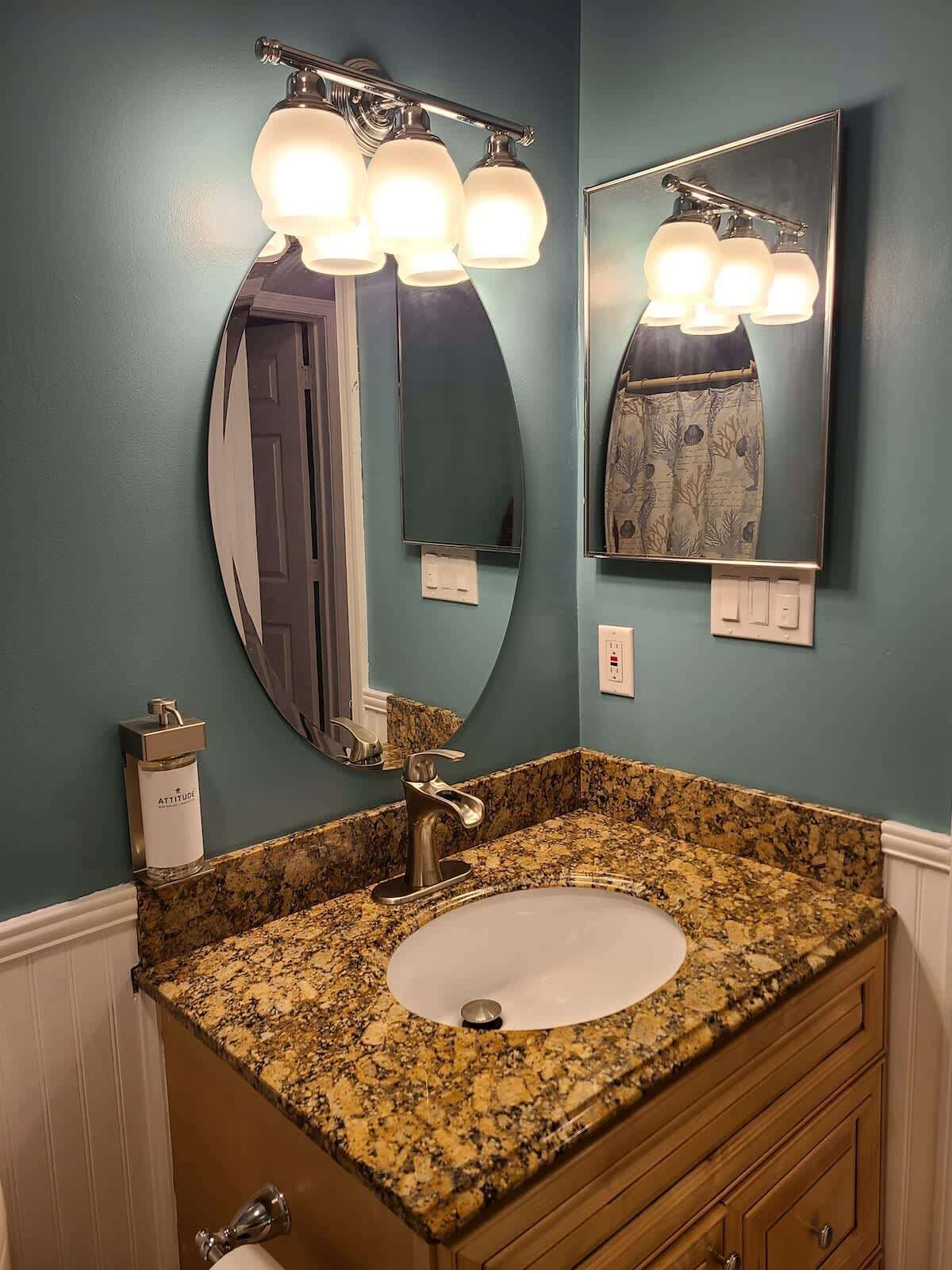 A bathroom with a granite countertop sink, a vanity with a mirror, and a wall-mounted light fixture.
