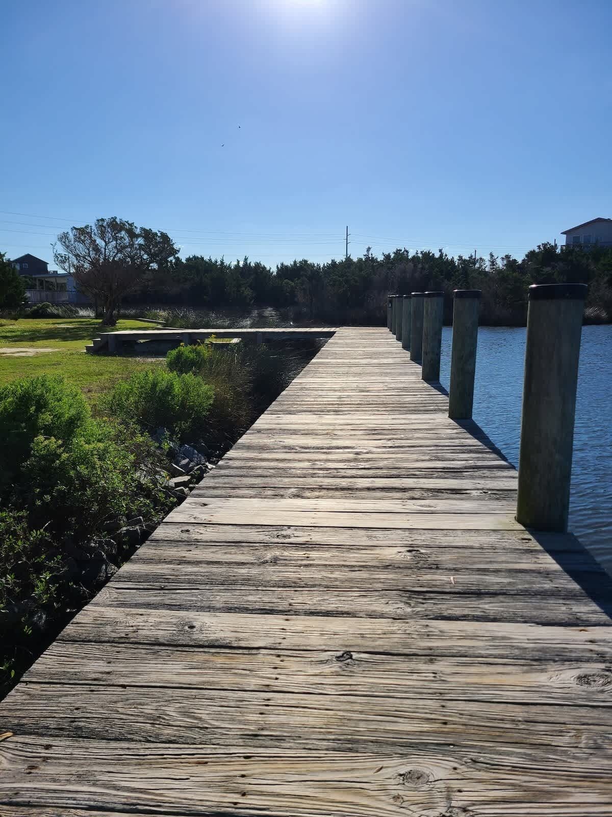 A wooden pier extending into a body of water, with a grassy area and trees on one side and a building on the other.