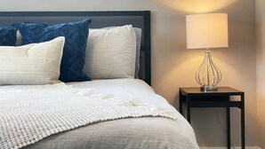 Un lit bien fait, agrémenté d’un coussin blanc et bleu, d’un plaid blanc tricoté et d’une lampe de chevet posée sur une petite table noire.