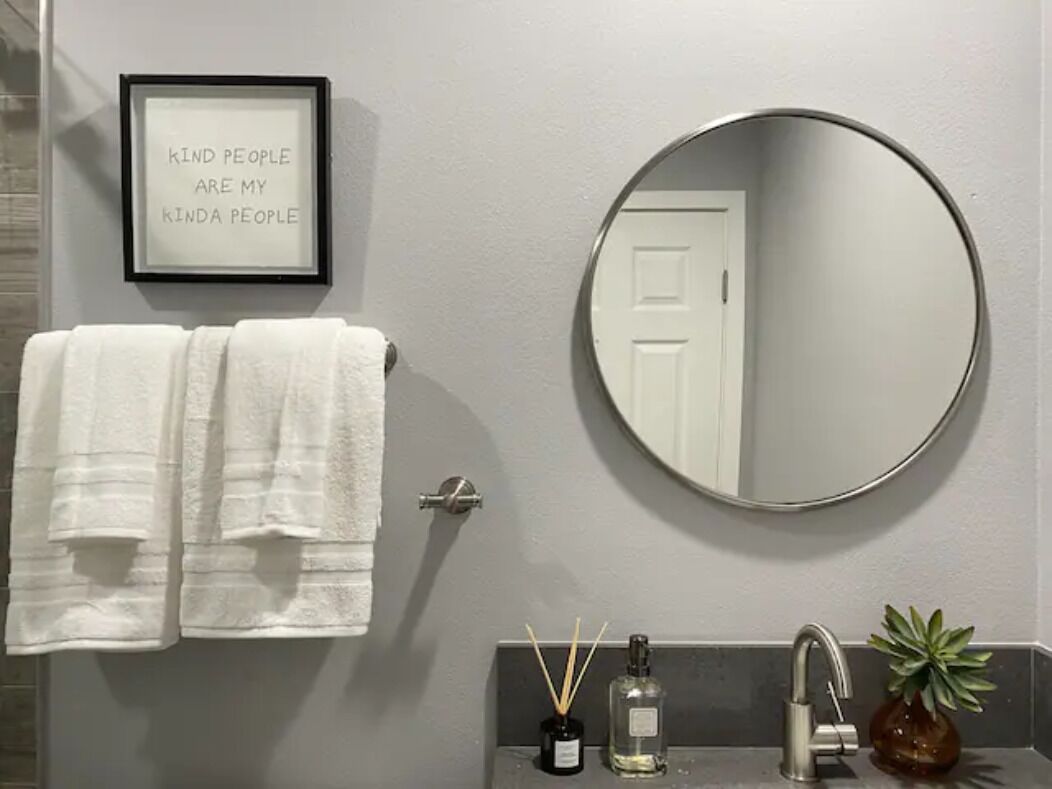 A bathroom with a framed quote on the wall, a round mirror, a towel rack with two towels, a sink with a bottle and a plant.