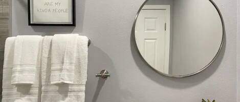 A bathroom with a framed quote on the wall, a round mirror, a towel rack with two towels, a sink with a bottle and a plant.