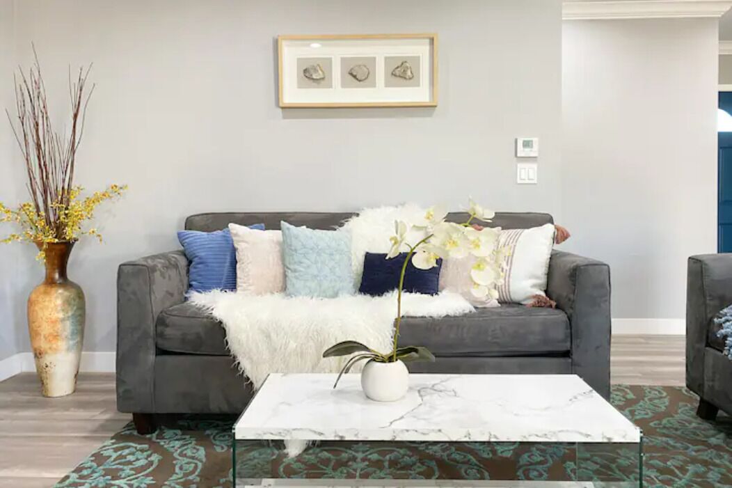 A living room with a grey sofa, white and blue cushions, a white fluffy throw, a vase with yellow flowers, and a framed picture on the wall.