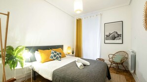 A neatly arranged bedroom with a bed, bedside lamps, a potted plant, a wicker chair, and a framed picture on the wall. - Spacious, family-friendly apartment right on Zurriola Beach (Donostia)