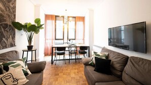 A living room with a sofa, a TV mounted on the wall, a dining area with a table and chairs, and a large plant. - Spacious, family-friendly apartment right on Zurriola Beach (Donostia)