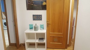 Un couloir avec une porte en bois, une étagère blanche sur laquelle se trouve une tablette, et un panneau indiquant « BONJOUR » au-dessus.