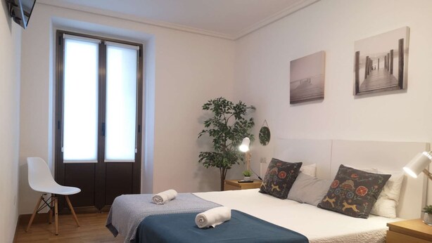 Une chambre à coucher avec un lit, deux oreillers, une table de chevet, une lampe, une chaise, une plante et une fenêtre avec des stores.