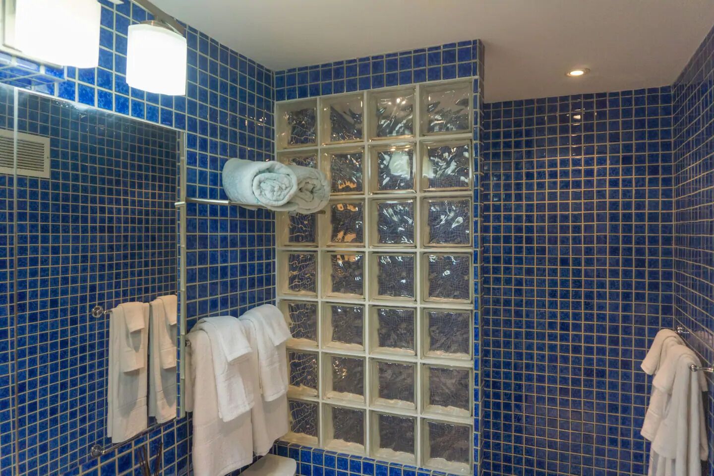 A bathroom with blue-tiled walls, a large window with frosted glass, and multiple white towels hanging on a rack.