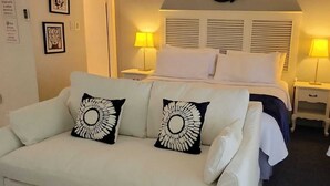 Et hotelværelse med en seng, en hvid sofa med pyntepuder, to sengeborde med lamper og et vindue med gardiner.
