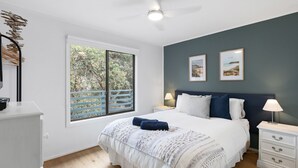 Un dormitorio con una cama grande, dos mesitas de noche con lámparas, un ventanal con vistas a árboles y dos cuadros en la pared.