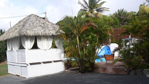 Una pequeña estructura con techo de paja y fachada blanca, con un balcón cubierto, junto a una zona de piscina con tobogán.