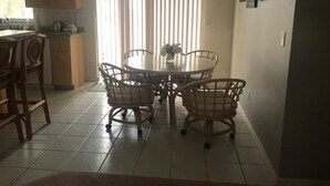 Uma área de refeições com uma mesa redonda e quatro cadeiras, cozinha ao fundo e uma janela com persianas.