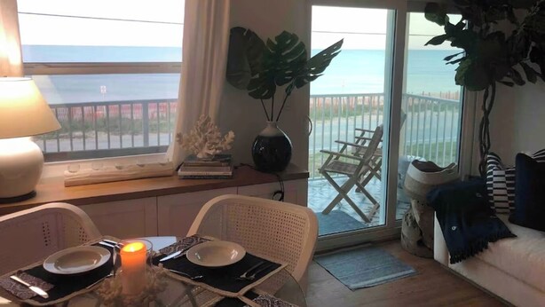 A dining area with a table set for two, a candle, and a view of the sea through a balcony door. - Luxurious Oceanfront Ormond Getaway (Ormond Beach)