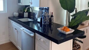A kitchen with white cabinets, a black countertop, and a sink. There is a plant on the counter and a basket underneath. - Luxurious Oceanfront Ormond Getaway (Ormond Beach)