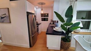 A compact kitchen with white cabinetry, a stainless steel refrigerator, a small dining area with a white chair, and a large green plant. - Luxurious Oceanfront Ormond Getaway (Ormond Beach)
