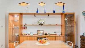 Ein Essbereich mit Holztisch, Stühlen und einer Obstschale in der Mitte. Darüber befinden sich Regale mit verschiedenen Gegenständen und ein Schrank mit Glastüren.