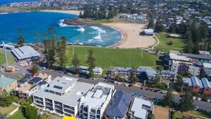 An aerial view of a coastal town with a beach, residential buildings, and a large hotel with multiple floors and a flat roof.