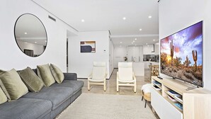 A modern living room with a grey sofa, a round mirror, a flat-screen TV, and a minimalist kitchen in the background.