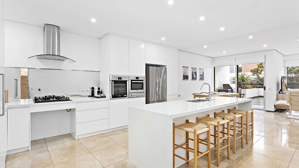 A modern kitchen with a central island, built-in ovens, a stainless steel range hood, and a large window overlooking a garden.