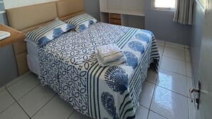 Un dormitorio con cama, mesita de noche, estante en la pared, ventana con cortinas e iluminación en el techo.