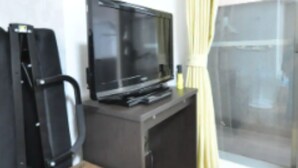 A room with a wooden table, a black TV on a stand, a black chair, and a window with yellow curtains. - IKminami6jo residence 101 (Sapporo)