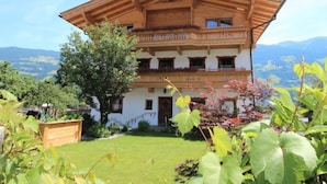 A two-story building with a sloped roof and balconies, surrounded by greenery and mountains in the background.