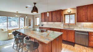 A modern kitchen with wooden cabinets, a large island with bar stools, and a view of the outdoors through large windows. - Riverfront Retreat in Nathrop: Fire Pits & Hot Tub (Nathrop)