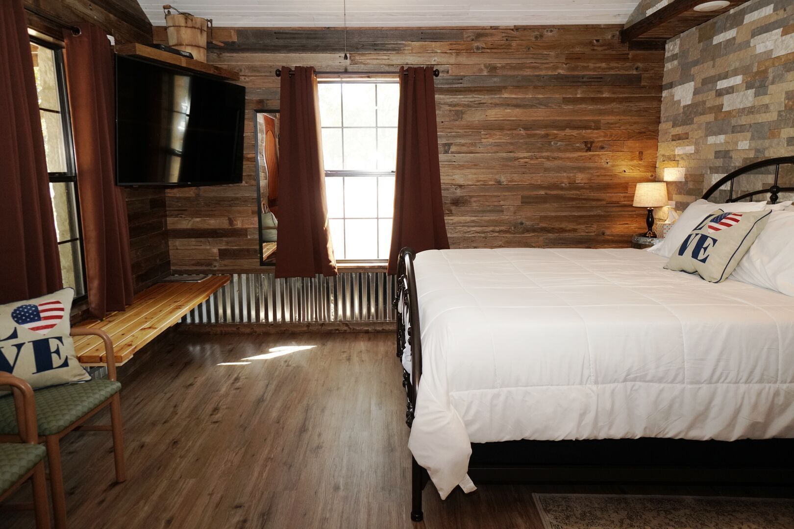 Un dormitorio con piso de madera, una cama con ropa de cama blanca, un televisor montado en la pared y una ventana con cortinas.